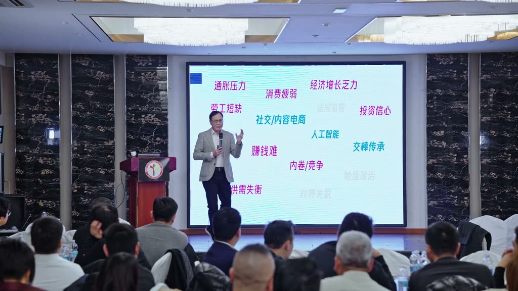杨国安教授手持麦克风站在大屏幕前讲课,屏幕上列着通胀压力、劳工短缺等词
